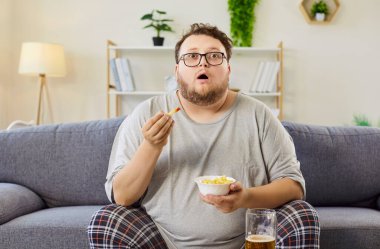 Overweight man breaks diet with fries, beer. He watches television on the sofa, snacking mindlessly and showing obesity and sedentary leisure. Unhealthy habits raise calories and risk.