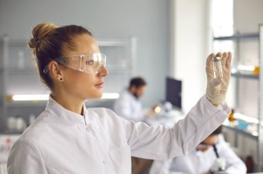 Young woman scientist looks at a test tube with white substance. A woman researcher in a white coat and goggles holds a test tube with white liquid