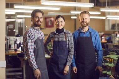 Portrait of three Caucasian experienced smiling shoe and garment factory workers at work. Two men and one woman dressed in work aprons stand in a large workshop and look into the camera