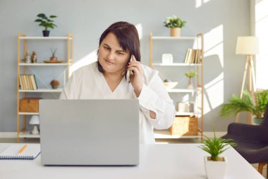 Plus size woman sitting at laptop and having phone conversation in office. Portrait of smiling overweight woman sitting at her desk in front of laptop computer and talking on phone