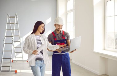 Woman discusses future house building with architect or engineer. Happy young girl with paper roll and man builder in safety hat and uniform look at construction plan on modern laptop computer device