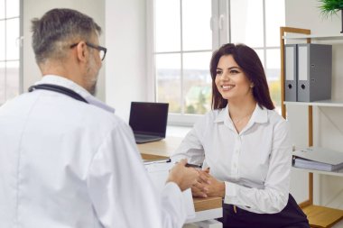 Happy, young brunette woman at appointment with doctor with glasses, who makes entries in the patients chart. Beautiful patient listens attentively and looks approvingly at her attending physician.
