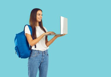 Smiling student girl with backpack in casual clothes working on laptop on blue background looking at screen. Education, learning, studying, work, student life concept. Wearing jeans and white t-shirt.