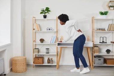 Young black woman feels severe back pain while at home in the living room. Pretty african american woman hardly moves, rubs tense back muscles, suffers from pain in the lumbar region of the kidneys.