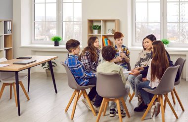 Group of elementary school children and their teacher discussing something while sitting in a circle on comfortable chairs in a modern classroom all together