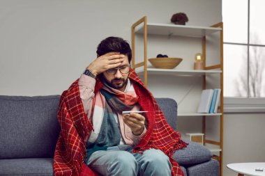 Sick man with fever looks confusedly at thermometer showing high body temperature. Worried young man wrapped in plaid grabs his head while sitting on sofa at home. Concept of viral infection.