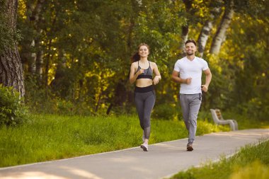 Smiling young man and woman jogging together along park path surrounded by green trees. Active couple enjoying morning run outdoors, maintaining healthy lifestyle, fitness routine and positive mood.