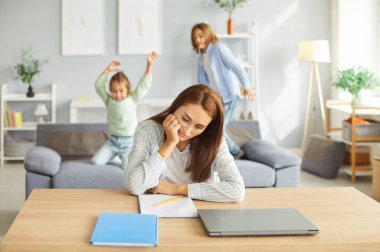 Portrait of exhausted upset mother sitting at the desk at home trying to work tired from her noisy children daughter and son playing on sofa in background. Problems of parenthood concept