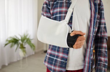 Male patient in support brace and white forearm scarf sling bandage immobilizer after surgery on his broken arm, elbow, hand, or wrist, cropped closeup shot. Accident, injury, and treatment concept