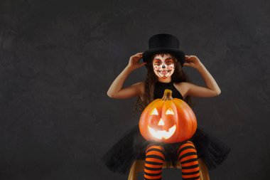 Child having fun on Halloween. Portrait of kid in spooky costume. Happy girl with beautiful skull makeup looking at camera sitting with cute smiley festive Jack-o-lantern on dark copyspace background