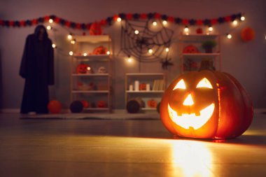 Festive background with beautiful smiley carved jack-o-lantern pumpkin with lit candle inside placed on wooden floor and lighting up dark festively decorated living room. Halloween celebration concept