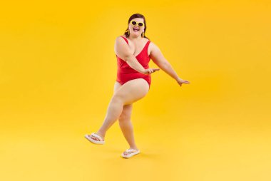 Portrait of overjoyed excited happy funny fat woman in red swimsuit and sunglasses having fun and dancing isolated on a studio yellow background. Summer holiday trip and vacation concept.