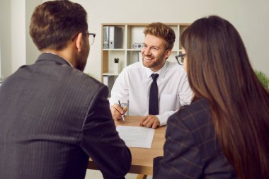 Friendly, polite real estate agent meeting with clients and offering them contract. Happy man sitting at office desk together with young couple, smiling and giving them pen and paper agreement to sign