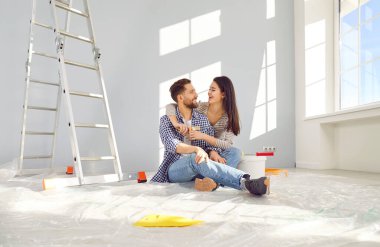 Smiling happy couple painting the wall of their new home. Married man and woman sitting on the floor hugging, looking at each other and planning repair renovation preparing to move into a new flat.