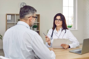 Young female doctor in white coat, glasses and stethoscope holding clipboard, asking questions and giving medical consultation to mature man patient while sitting at her desk with laptop at hospital
