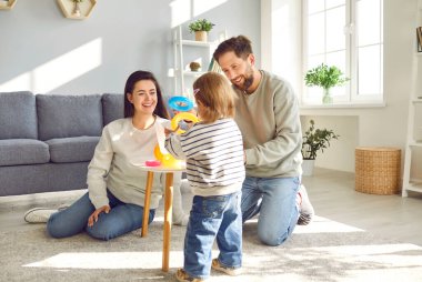 Young mother, father and their little daughter spend time at home in cozy childrens room. Parents are sitting on floor and playing with different toys with their child. Family holiday concept.