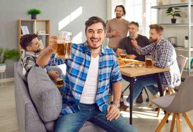 Portrait of attractive man holding a glass of beer looking at camera and smiling with a company of cheerful happy male friends talking and drinking beer in background. Men enjoying party together.