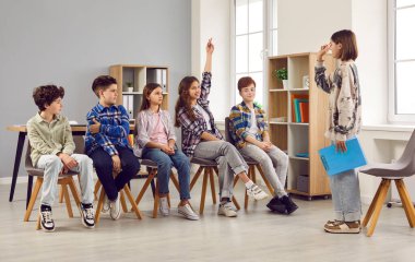 Group of school children sitting in a row in the class room and answering questions of a peer student girl who acts as a teacher. Back to school, education, discussion concept