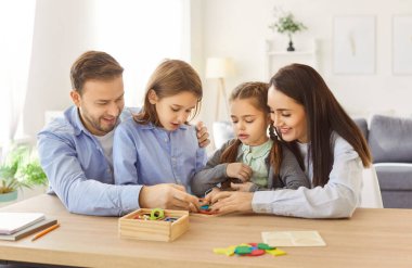 Portrait of children collecting wooden colorful geometric figures with mother and father sitting at table in living room at home. Young happy family playing in educational game with kids together.
