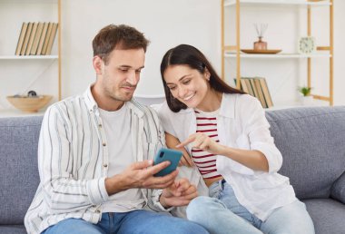Cheerful family couple sits on a couch at home, enjoying online entertainment in internet together on a mobile phone. They laughing, talking, and bond during leisure time in a connected environment.