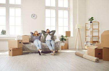 Purchase of new housing. Satisfied young married couple relaxing and enjoying sitting on sofa in new home. Husband and wife sitting on sofa with their hands behind their heads in room between boxes.