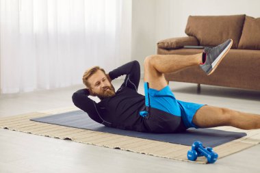Strong motivated man doing an abs workout on an exercise mat at home. Handsome young Caucasian guy in modern sportswear doing bicycle crunches lying on a fitness mat in the living room