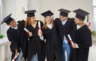 Team of university students celebrating graduation and having fun together. Group of happy cheerful elated multiethnic graduates and friends in hats and robes holding diplomas and hugging in classroom
