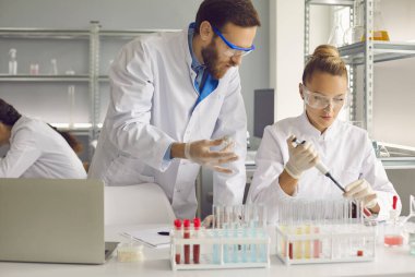 Young medical staff in special clothes are conducting research, a man leader helps a woman, holding an empty test tube. A woman fills an empty test tube.