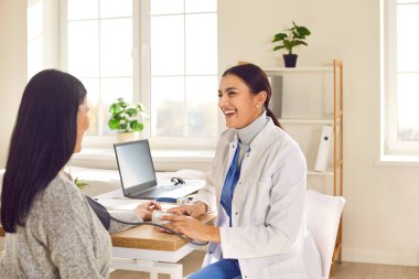 Portrait of a smiling attractive woman doctor therapist measuring blood pressure of a young female patient sitting back in the office in clinic during medical examination. Healthcare concept.