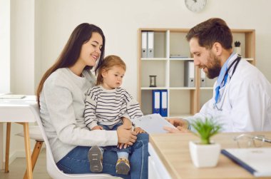Young mother with small child listens to male pediatrician who fills out medical form and prescribes treatment. Smiling woman with two-year-old daughter at doctors appointment in clinic.