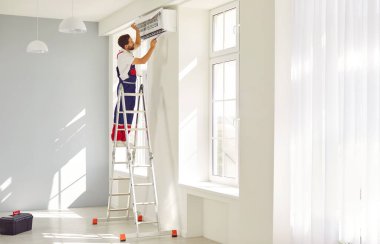 Profesyonel erkek teknisyen yeni eve modern duvar kliması kuruyor. İş üniformalı bir adam parlak, ferah bir odada merdivenin üzerinde duruyor ve klimayı tornavidayla monte ediyor..