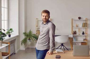 Portrait of a young businessman at home. Young business man in a casual shirt and jeans standing by his working desk with a laptop, book, and smartphone in a modern room interior