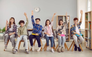 Happy school students sitting in row and raising their hands to answer. Group of smiling elementary schoolchildren in casual clothes answering question in classroom at school