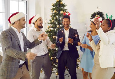 Happy group multiracial friends in Santa hats men and woman celebrating Christmas talking with sparklers in hands near Xmas Tree at home or modern office. Celebrate winter holidays, New Year concept.