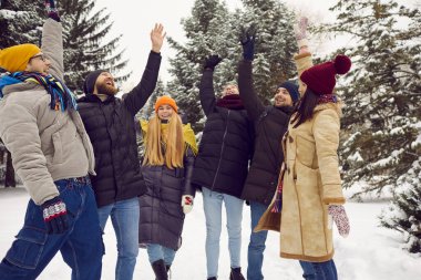 Team of happy millennial friends all together having some outdoor fun in snowy winter park. Group of young people in warm hats, coats and jackets standing in circle among trees and joining their hands