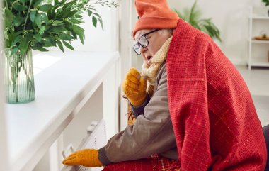 Oturma odasında oturan yaşlı bir adamın portresi battaniyeye sarılı ve kış şapkası elektrikli ısıtıcıda ısınmaya çalışıyor. Evde donan olgun bir adam. Isıtma sorunları konsepti.