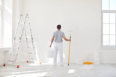 Portrait of young man painter from repair service in uniform standing back in empty room and holding a bucket with paint roller going painting a wall. Renovation and renovating house concept.