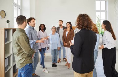 Business team meeting in their office workplace and having a discussion. Group of different happy diverse young and senior people standing in the office and listening to a young male speaker