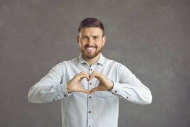 Handsome caucasian young man expresses a gesture of love while standing on a gray background. Smiling man in front of the camera shows his heart with his hands. Concept of romance and human emotions.