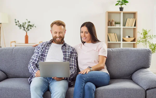 Happy family couple using a laptop together. Married man and woman sitting on the couch at home, using a notebook computer, surfing the net, shopping at an online marketplace, or watching a movie