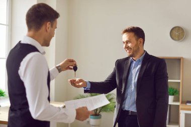 Portrait of professional man realtor agent standing in office with signed contract and giving house key to a happy smiling man customer and new homeowner. Real estate market or car purchase concept.