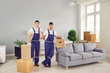 Moving service. Portrait of two friendly workers of moving and delivery company who transport cardboard boxes. Male loaders in overalls standing in room with cardboard boxes and smiling at camera. 