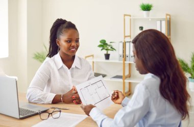 Masasında dizüstü bilgisayarı, tasarımcısı, mimar bürosu danışmanlığı olan genç Afro-Amerikan kadınları var. Kadın iş arkadaşı ev planı, inşa, satın alma ve satma maliyetleri, gayrimenkul iş hizmetleri