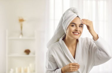 Beautiful woman in morning after shower before applying makeup cleanses skin using cotton pad. Smiling girl in bathrobe and with towel on head in white interior. Concept of face cleaning and skin care