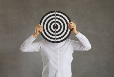 Woman hiding her face behind a shooting target standing on a grey studio background. Concept of setting an objective, reaching business goal, hitting target audience and achieving success