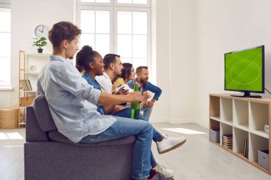 Multiracial friends young guys and girls watching soccer football game on TV together sitting on couch in living room. Football fans closely following progress of game. Sport fans, friendship concept.