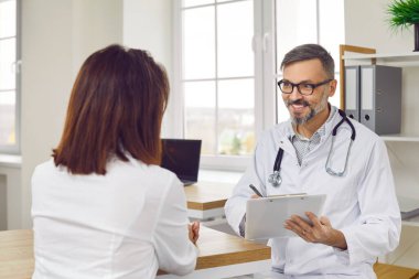 Friendly positive male doctor writing prescription or making notes in patients card during medical consultation. Smiling middle-aged man consults female patient at workplace in medical office.