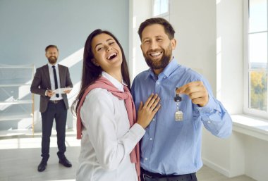 Happy couple embracing showing new house keys to camera. Real estate agent and married couple standing in empty room of new apartment, excited by moving. Buying new house concept