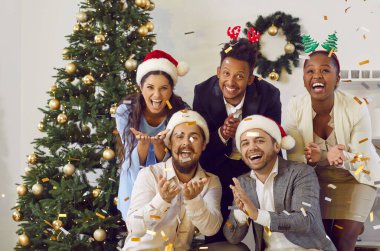 Happy cheerful multiethnic friends catching confetti on New Year party near Christmas tree at home. They smiling laughing looking at camera. Christmas spirit, celebrating winter holidays concept.