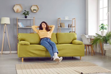 Happy relaxed young woman sitting on comfortable sofa. Beautiful serene girl in casual clothes holding hands behind head resting on couch at home enjoying free time, lazy weekend time
